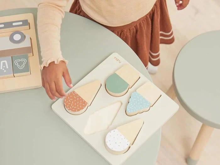 En barnehånd rækker ud efter et is-puslespil af træ på et bord med farverige isformede brikker og matchende åbninger.