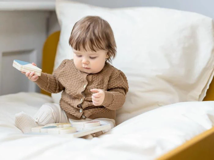 En baby i en brun sweater sidder på en seng og leger med træklodser. Babyen kigger ned, fokuseret på legetøjet, omgivet af hvidt sengetøj.