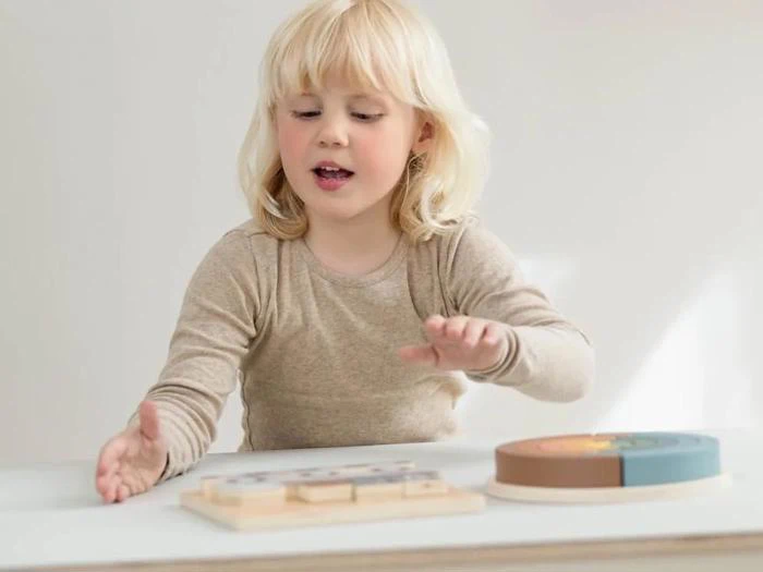Et lille barn med blondt hår leger med et træpuslespil ved et hvidt bord og rækker interesseret og fokuseret frem.