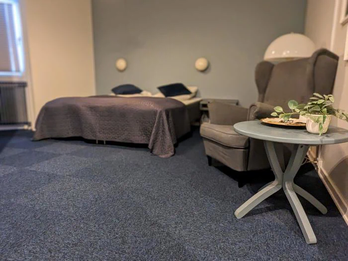 Et hyggeligt soveværelse med en dobbeltseng, gråt sengetøj, en lænestol og et rundt sidebord med en plante og en bakke på, sat op mod blåt gulvtæppe og blød belysning.