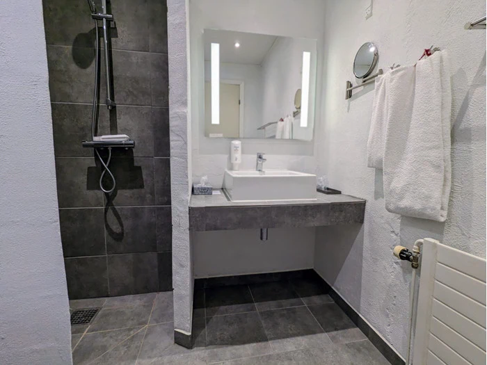 Moderne badeværelse med bruseniche, flydende håndvask under et stort spejl, hvide håndklæder og mørkegrå gulvfliser.