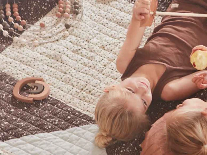 To små børn ligger på en mønstret dyne, den ene holder en pind og den anden et æble. Trælegetøj og en ting med perler ligger spredt i nærheden.