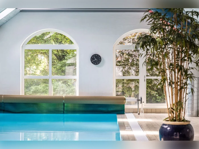 Indendørs swimmingpool med klart blåt vand, store buede vinduer med udsigt til grønne træer udenfor, et ur på væggen og en potteplante i højre side.