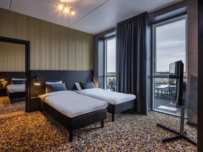 Moderne hotelværelse med to enkeltsenge, et stort vindue med udsigt over byen, mørke gardiner, et tv på en stander, et vægspejl og et mønstret gulvtæppe.