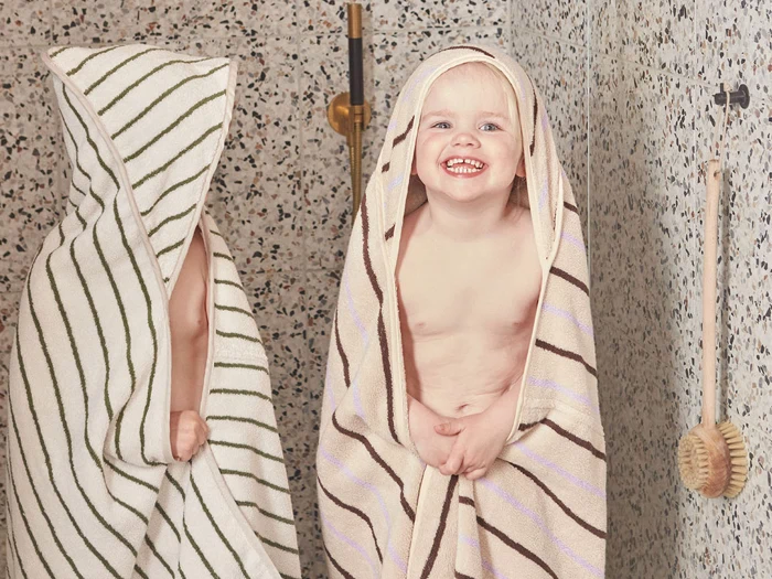 To små børn indhyllet i stribede håndklæder står i et flisebadeværelse. Det ene barn smiler til kameraet, mens det andet for det meste er dækket af sit håndklæde. En badebørste af træ hænger på væggen