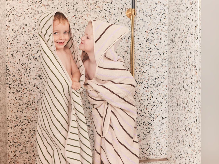 To små børn indhyllet i stribede håndklæder står og smiler i et flisebelagt bruseområde. Det ene barn smiler, mens det andet ser ud til at læne sig legende frem for at kysse eller hviske.