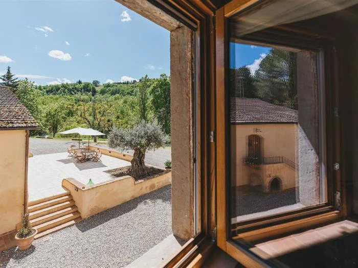 Udsigt fra et åbent vindue, der viser en solrig gårdhave med bord, stole, parasol, stentrapper, træer og en bygning med balkon i baggrunden.