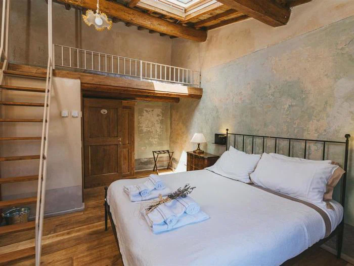 Rustikt soveværelse med dobbeltseng, hvidt sengetøj, synlige træbjælker, hems med stige, trægulv og ovenlysvindue. Der ligger håndklæder og lavendel på sengen.