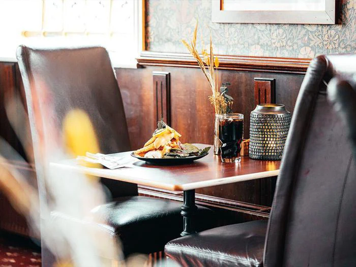 Et hyggeligt restaurantbord med to læderstole, en tallerken med mad, et glas sodavand og dekorative tørrede planter i en vase på en træoverflade.