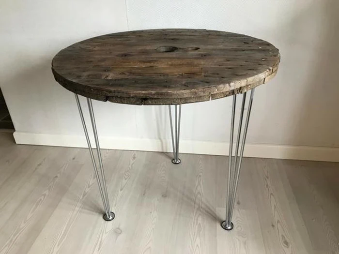 Et rundt træbord med en rustik bordplade og fire tynde hårnåleben i metal står på et lyst trægulv.