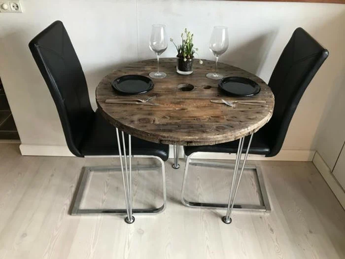 Et lille rundt træbord med to sorte stole, dækket til to med tallerkener, vinglas, bestik og en lille plante i midten.