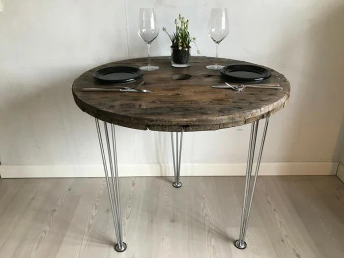 Et rundt træbord med hårnåleben i metal, dækket til to med sorte tallerkener, sølvtøj, vinglas og en lille potteplante i midten.