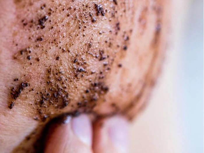 Et nærbillede af en persons hud, der bliver eksfolieret med en grynet, brun scrub, sandsynligvis en kaffe- eller sukkerscrub, som gnides ind med fingrene.