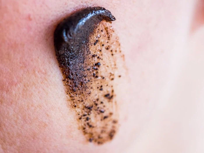 Et nærbillede af en persons kind med en klat ansigtsscrub, der indeholder mørke eksfolierende partikler.