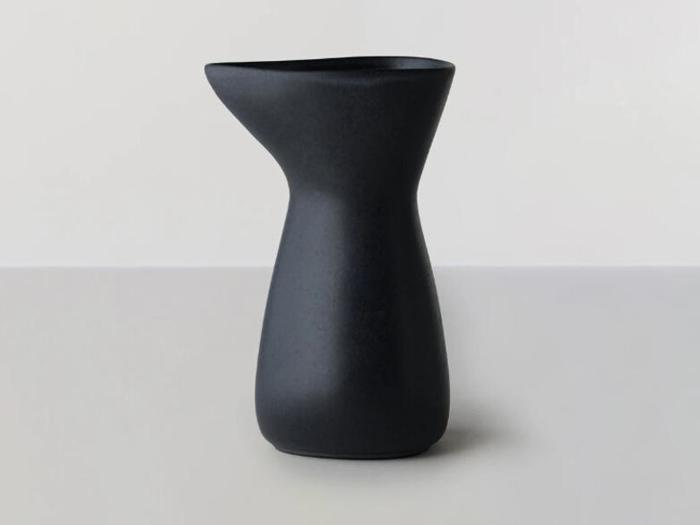 En matsort keramisk kande med et asymmetrisk, moderne design står på en lysegrå overflade mod en lys baggrund.