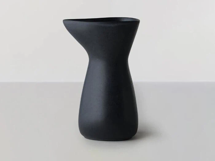 En matsort keramisk kande med et asymmetrisk, moderne design står på en lysegrå overflade mod en lys baggrund.