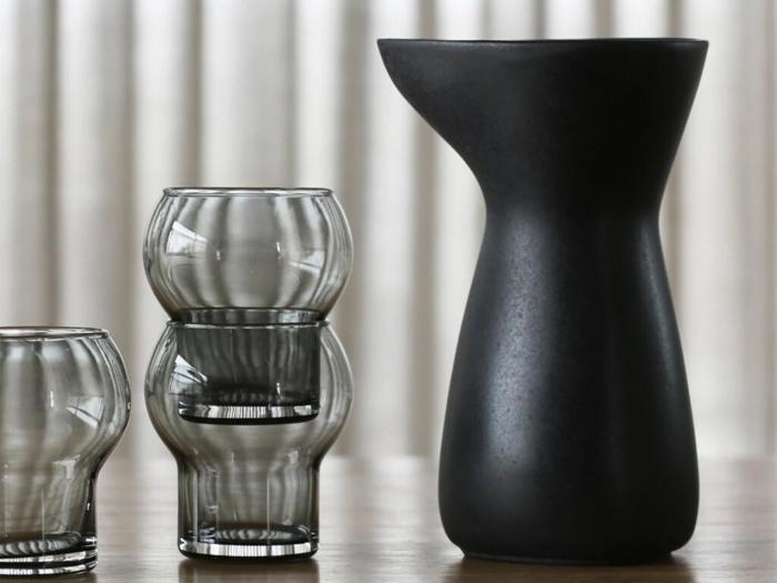 En matsort kande står ved siden af to stablede, afrundede klare glas på en træoverflade med lodrette gardiner i baggrunden.