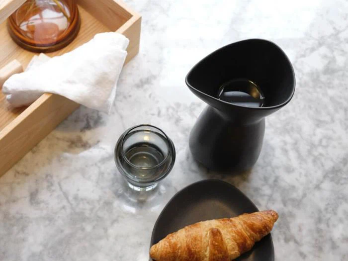 En croissant på en sort tallerken, en lille sort kande, et glas vand og en træbakke med en hvid serviet på en marmoroverflade.