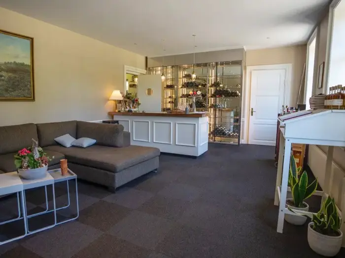 En hyggelig lounge med en sektionssofa, sofaborde med planter, en bar med glashylder og store vinduer, der lukker naturligt lys ind.