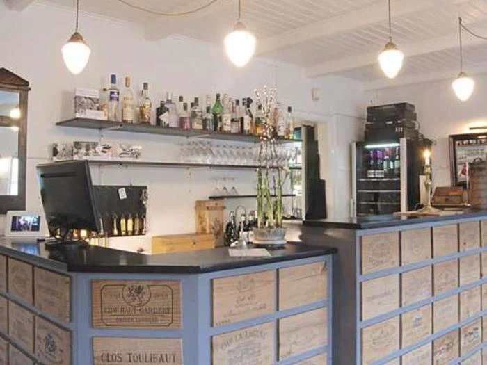 En bardisk med skuffer dekoreret med vinkassetryk, flasker og glas på hylder, en computerskærm og pendellamper, der hænger ned fra loftet.