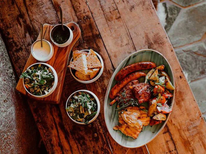 Et træbord med grillet kød, pølser, ristede grøntsager, brødskiver, to små salater og to dypsaucer i skåle.