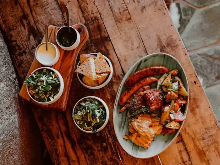 Et træbord med grillet kød, pølser, ristede grøntsager, brødskiver, salater og dypsaucer arrangeret i skåle og på tallerkener.