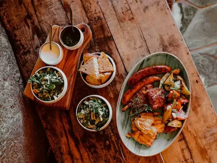 Et rustikt træbord dækket med grillet kød, pølser, ristede grøntsager, to salater, brød og saucer i små skåle.