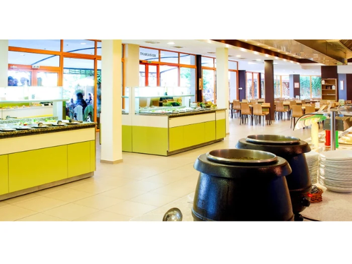 Lyst, moderne cafeteria med gule buffeter, suppegryder og stabler af tallerkener; borde og stole er placeret nær store vinduer i baggrunden.