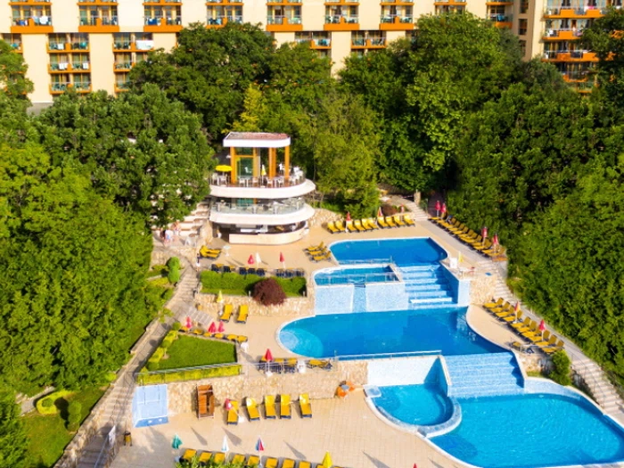 Luftfoto af et hotels poolområde med flere pools, liggestole, parasoller, grønne områder og en poolbygning i flere niveauer med en hotelbygning i baggrunden.