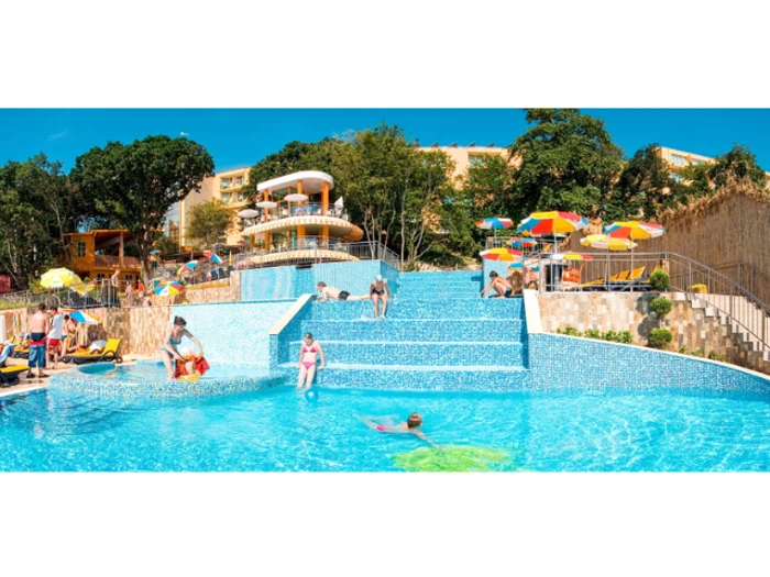 Folk svømmer og slapper af i en stor udendørs pool i flere niveauer omgivet af træer, farverige parasoller og liggestole under en lyseblå himmel.