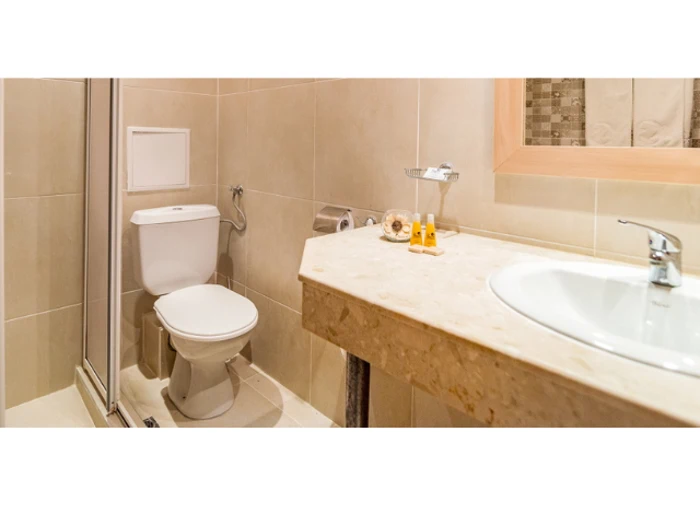 Moderne badeværelse med beige fliser, toilet, glasbruser, væghængt toiletpapirholder, marmorvask, toiletartikler og et vægspejl over vasken.