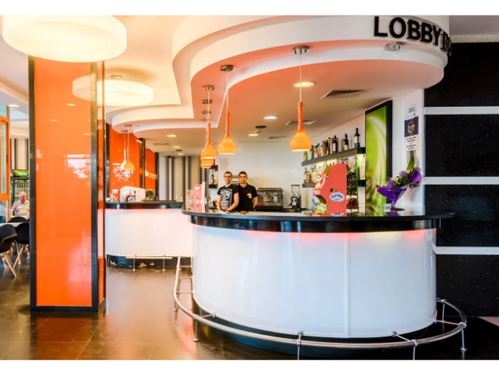 En moderne, stærkt oplyst lobbybar med to personer bag den buede disk, orange detaljer og flasker på hylderne.