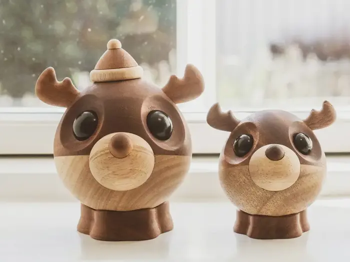 To runde træfigurer med store sorte øjne og gevirer, som ligner rensdyr, sidder på en hvid flade foran et lyst vindue.