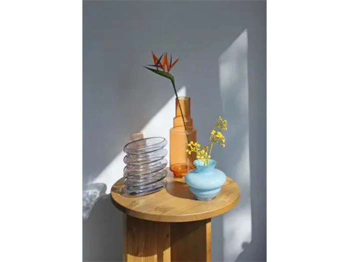 Et rundt træbord med glasvaser, en med en paradisfugl, en anden med gule blomster og en klar spiralformet vase, op ad en grå væg med sollys og skygger.