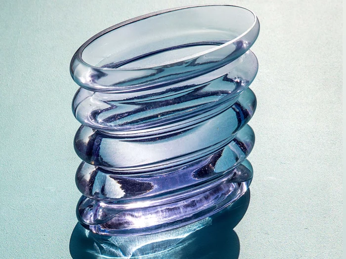 En bølget, transparent, lilla glasvase står på en struktureret lyseblå overflade og kaster bløde skygger og refleksioner.