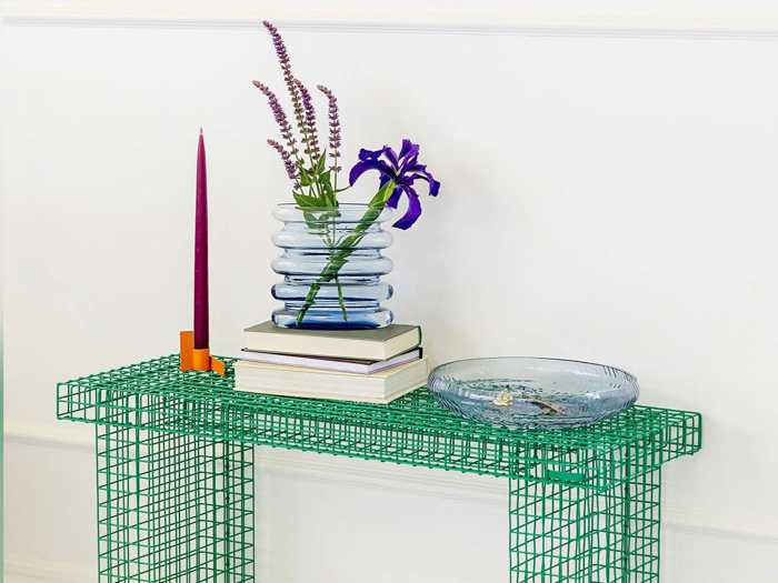 På et grønt trådbord står et lilla stearinlys, stablede bøger, en blå vase med blomster og en klar glasskål op ad en hvid væg.