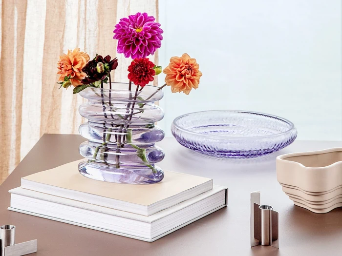 En glasvase med farverige georginer står på stablede bøger ved siden af et rundt glasfad og en beige skål på et lysebrunt bord ved et vindue med gennemsigtige gardiner.