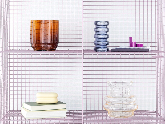 På en lyserød trådhylde står ravfarvede og klare vaser, stablede bøger, et hvidt stearinlys, blå glasdekorationer og små lilla kubiske genstande arrangeret pænt.