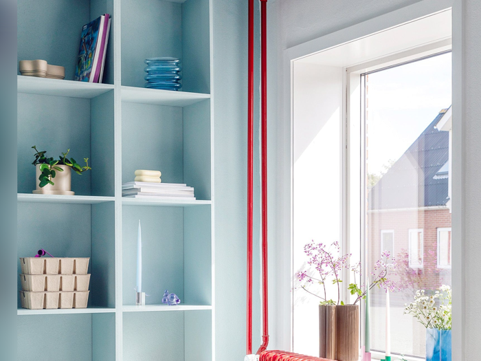 Lyseblå hylder med forskellige ting, bl.a. planter, bøger og tallerkener, ved siden af et solbeskinnet vindue med blomster i vaser og udsigt til huse udenfor.