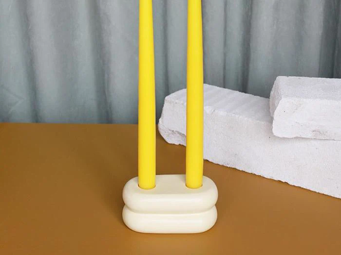 To høje gule stearinlys står i en afrundet cremefarvet lysestage på en sennepsfarvet overflade med hvide strukturblokke og grå gardiner i baggrunden.