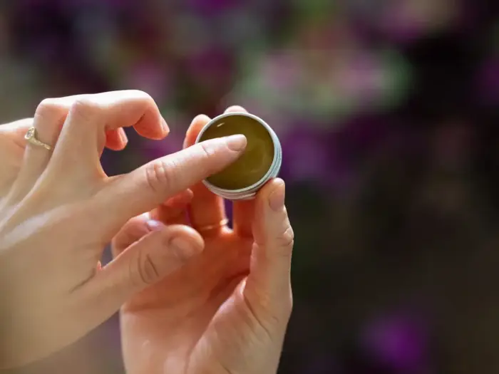 En person dypper sin finger i en lille dåse med grønlig balsam med en sløret lilla og grøn baggrund.