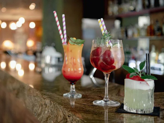 Tre farverige cocktails med stribede sugerør og pynt står på en bardisk med en sløret baggrund af flasker og varm belysning.