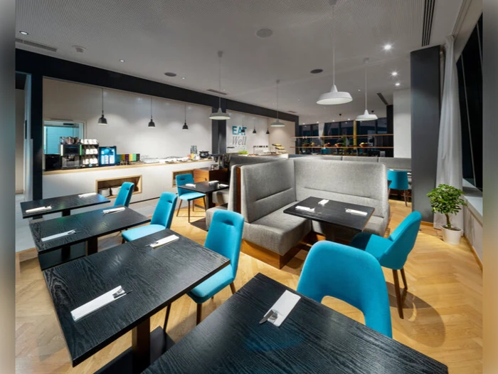 Moderne spiseplads med mørke træborde, blå stole, grå siddepladser og pendellamper. Buffetopstilling og kaffemaskiner ses i baggrunden; potteplante i hjørnet.