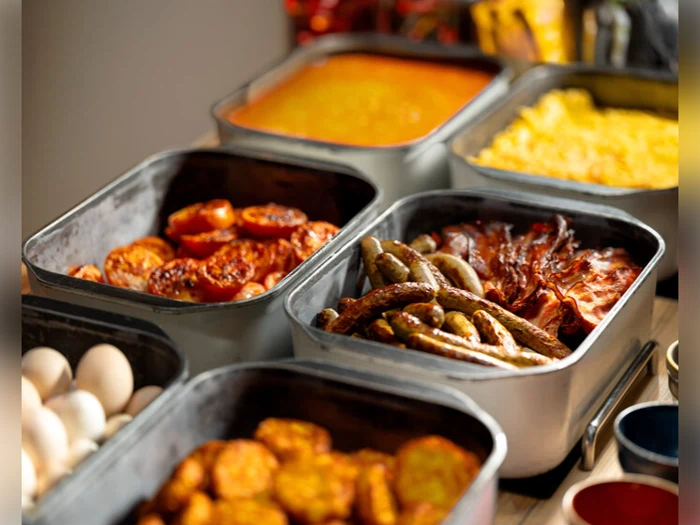 Metalbakker fyldt med morgenmad, herunder pølser, bacon, hash browns, baked beans, røræg, æg og ristede tomater, stillet frem som en buffet.