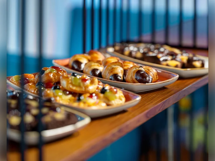 Tallerkener fyldt med forskelligt bagværk, bl.a. wienerbrød med frugtovertræk, chokoladecroissanter og dessertstænger, anbragt på en træhylde bag metalstænger.