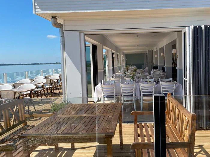 Udendørs siddeområde med træborde og -stole ved siden af en glasvæg, der fører til en indendørs spisestue med hvide stole og duge med udsigt over et vandløb.