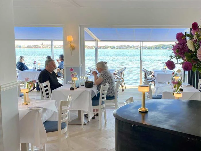 En restaurant ved vandet med par, der spiser ved borde med hvide duge nær store vinduer med udsigt over et roligt vandområde. Lyserøde blomster og blød belysning dekorerer scenen.