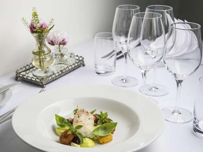 En gourmetret med fisk, grøntsager og krydderurter på en hvid tallerken, dækket på et elegant bord med tomme vinglas, vandglas, servietter og en lille blomsteropsats.