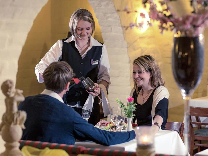 En tjener skænker vin til et smilende par, der sidder ved et bord i en hyggelig, varmt oplyst restaurant.