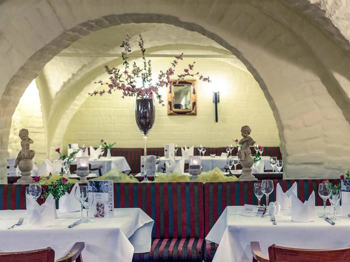 En hyggelig restaurant med buede lofter, hvide duge, pænt opstillede borde, elegante glas og en høj vase med lyserøde blomster som midtpunkt.